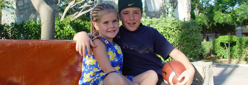 Molly and Jimmy sitting next to each other on a bench in Cabo San Lucas, Mexico. Molly has corn rows and is wearing a blue and yellow flowered dress. Jimmy is holding a football and has his right arm around Molly. He's wearing a blue t-shirt and khaki shorts