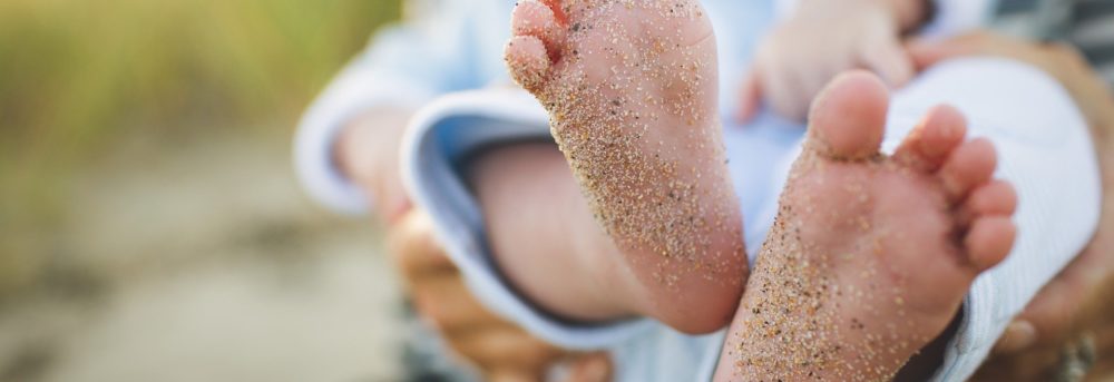 Baby's feet covered in sand. The baby is wearing a blue outfit. The background is greenish and blurry.