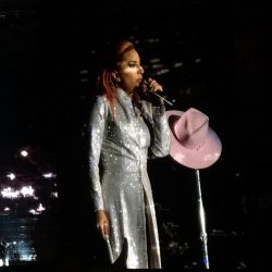 Lady Gaga wearing a long white coat holding a microphone with her pink hat sitting on the microphone stand in front of her