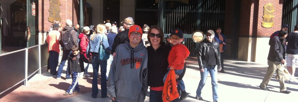 Jimmy, Shannon and Theo at AT&T Park. Jimmy is wearing an orange SF Giants cap, a gray Giants' sweatshirt and blue jeans. Shannon is wearing sunglasses, a dark shirt and dark pants. Theo is wearing a Giants' cap, an orange Giants' sweatshirt and carrying an orange Giants towel
