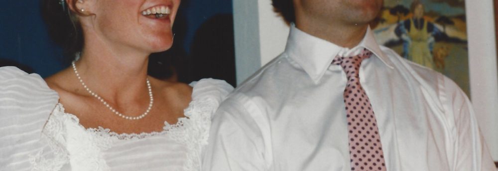 Dan and Margo on their wedding day. Margo is on the left wearing a white gown with pink roses and white flowers in her hair; Dan is wearing a white shirt and pink tie.