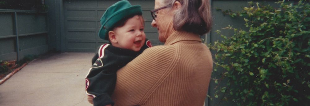 Mom holding Jimmy age 18 months. Mom is wearing glasses and a tan sweater; her gray hair is held back by a clip. Jimmy is wearing a green jacket and green hat.