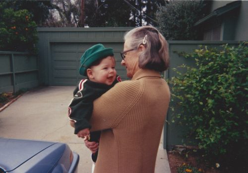 Mom holding Jimmy age 18 months. Mom is wearing glasses and a tan sweater; her gray hair is held back by a clip. Jimmy is wearing a green jacket and green hat.