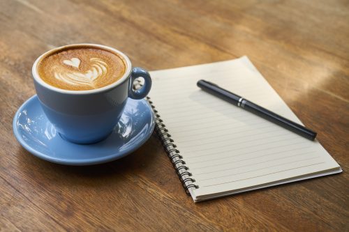 Latte in a blue tea cup sitting on a blue saucer with a heart drawn in the milk foam next to a spiral bound notebook with a black pen sitting on top. Both are sitting on a wooden table.