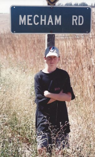 Sean is leaning against a sign that says "Mecham Road". He's wearing a baseball cap, black shirt and black shorts. His arms are crossed.