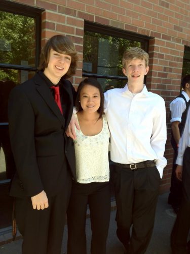 Niko and two friends at school. Niko is on the left wearing a black suit and red tie. His female friend is in the middle wearing a yellow top and black pants. His male friend is on the right wearing a white shirt and black pants.