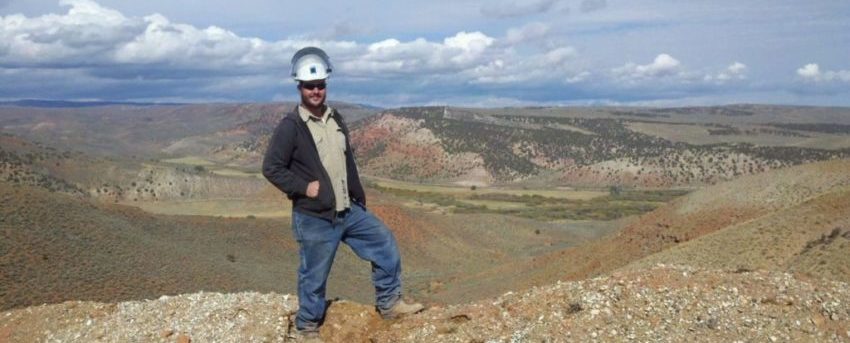 The author's son Jason wearing a white hardhat, dark jacket, light shirt and blue jeans standing on a hill in front of the pipeline