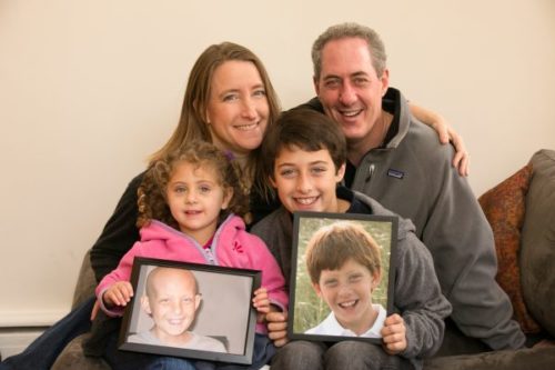 Nancy and her husband Michael are sitting behind their children Sarah and Ben. Nancy has her arm around Michael. Sarah is sitting on Nancy's lap wearing a pink jacket and holding a photo of Jacob who has a bald head from treatment. Ben is sitting on Michael's lap, searing a gray jacket, holding a photo of Jacob pre-diagnosis who has a full head of red hair.