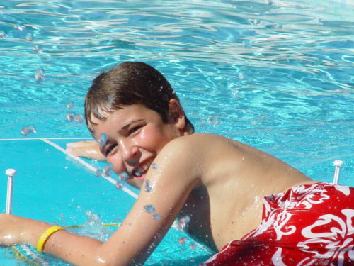 Jimmy at age 12 lying on a boogie board in a pool looking over his left shoulder at the camera wearing red and white swim trunks and a yellow LIVESTRONG band