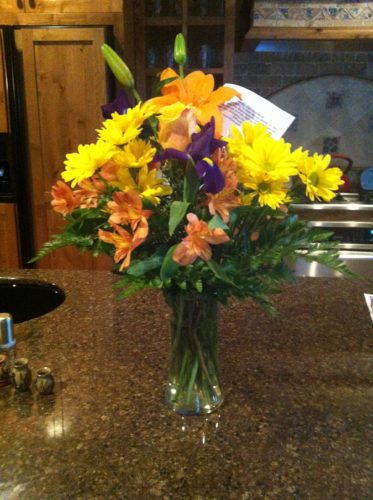 A bouquet of yellow and orange flowers with one purple flower and a card in a glass vase sitting on a brown marble kitchen countertop