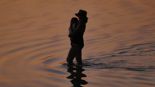 Silhouette of mom with a hat on and the child she's carrying. They're standing in water in fading light during sunset