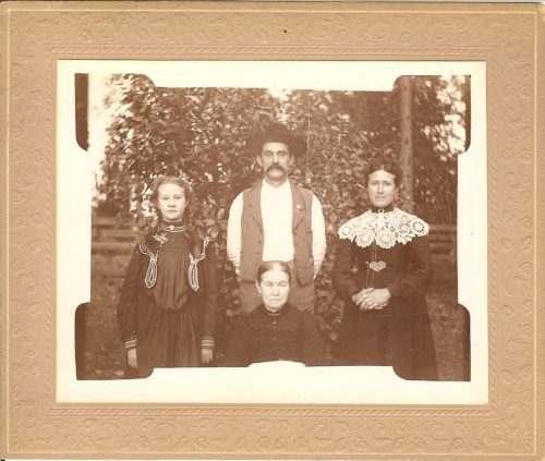 Mildred, James and Emily's daughter is standing on the left; James is standing in the middle with a mustache, wearing a hat; Jane Kissell, Emily's mom is seated in front of James and Emily is on the right. Sepia photo.