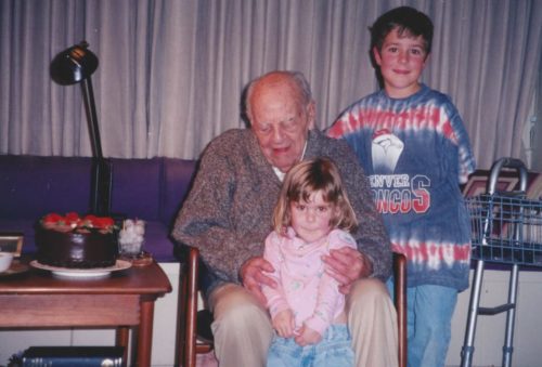 Dad's 100th birthday. On the left is his birthday cake. Dad is sitting in a chair with Molly standing between his legs. Jimmy is standing on the right wearing a Denver Broncos shirt