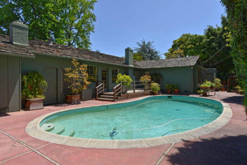 Backyard of Hurst Avenue. The pool, turquoise in color and kidney bean in shape, takes up most of the picture with the small wooden deck in the back and the green house behind the deck.