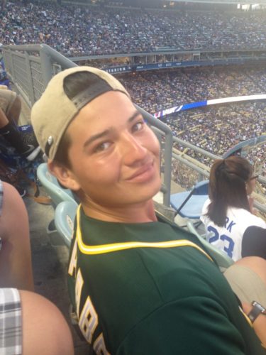 Tommy at a baseball game wearing an As jersey and a backwards baseball cap