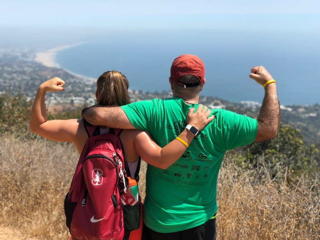 Molly and Dan standing with their backs to the camera overlooking the ocean near Santa Monica. Molly is making a muscle with her left arm and carrying a red Nike Stanford backpack. Dan is wearing a green shirt and red baseball cap and is making a muscle with his right arm.
