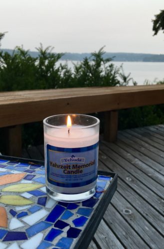 Lit yahrzheit candle for Katie sitting on blue, white and dark blue tile table on Karen's deck with view of the water beyond the low wall of the deck.