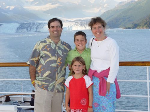 Dan wearing a Hawaiian shirt and tan pants on the left with Jimmy in the middle wearing a green shirt, Molly in front of him wearing a red shirt with white sleeves and Margo on the right wearing a white t-shirt with blue drawstring pants and a pink sweater tied around her waist all standing at the railing of a cruise ship in Alaska