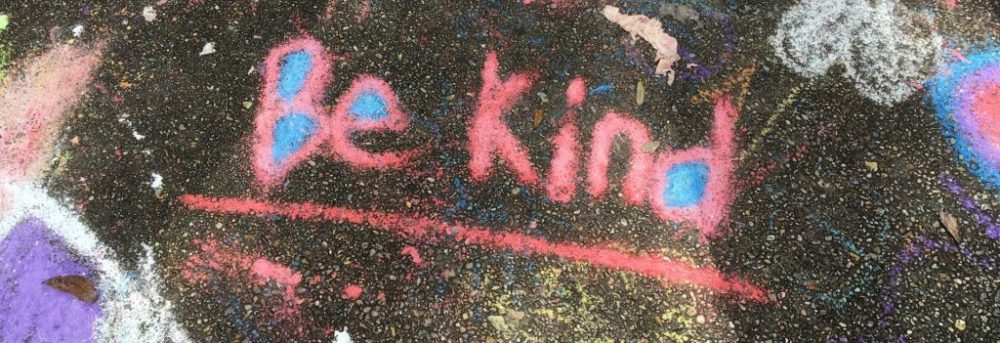 The words "Be kind" written in red chalk with the "B", "e" and "d" filled in with blue chalk. All on a black background.