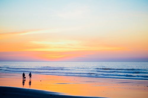 Two people walking at the water line on a beach. The sun is setting the sky is blue, yellow and orange and is reflected in the wet sand.