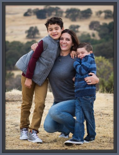Shani with her sons. Her older son is standing to her right with his left arm around Shani. He's wearing a long sleeve mulberry shirt with a light blue no sleeve down vest and tan pants. Shani is kneeling with her arms around both boys. She's wearing jeans and a long sleeve steel gray t-shirt. Her younger son is wearing a light and dark blue striped rugby shirt and jeans.