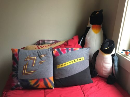 JF logo and JimmySTRONG on two throw pillow on Jimmy's bed which is covered with a red comforter. To the right of the pillows are two penguins, one taller than the other