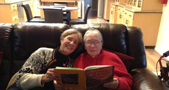 Diane and her mom are sitting on a brown leather couch in the living room, each holding the side of an open book. Diane is on the left wearing a black and white Christmas sweater. Diane's mom is on the right wearing a red sweatshirt.