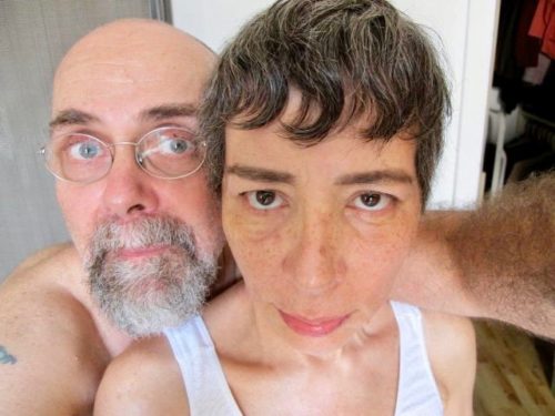 Donna is in front with short dark hair laced with gray, wearing a white tank top. Mark is behind her wearing glass and with a gray beard and mustache. He's wearing glasses