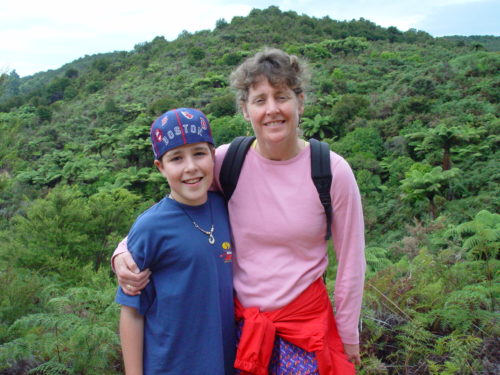 Jimmy and Margo on a hike in New Zealand standing in front of hill covered with green trees and bushes. Jimmy is on the left wearing a Boston Red Sox hat backwards and a blue t-shirt with the roadrunner and Tweety on it. Margo has her right arm around Jimmy. She's wearing a long sleeve pink shirt with a red jacket tied around her waist and a backpack on her back.