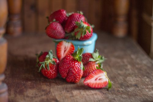 Strawberries with their stems on overflowing out of a small blue bowl. The bowl is on a wood table with four whole strawberries and two half strawberries in front of it.