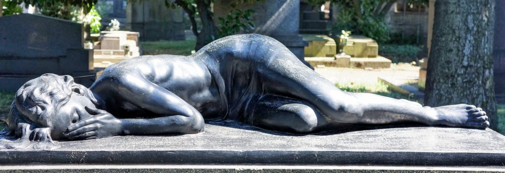 Statue of a woman lying on her side facing the camera. Her knees are bent and her right hand is covering her face as she grieves