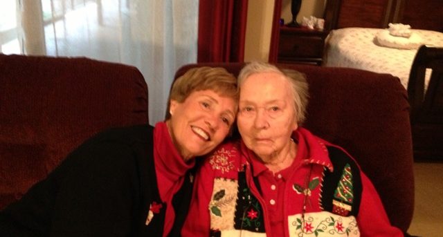 Diane and her mother sitting next to each on brown chairs in front of a window with sheer curtains. Diane is wearing a red turtleneck and black sweater. Diane's mom is wearing a red collar shirt and a Christmas vest