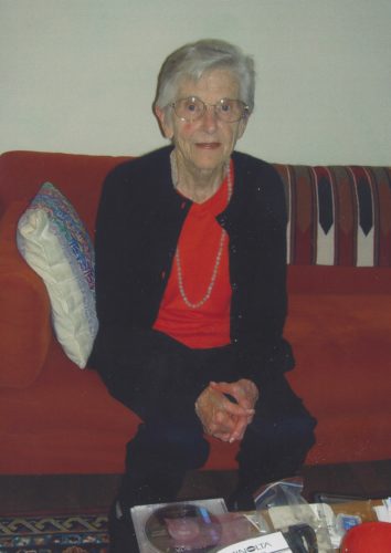 Mom in her early 80s sitting on a couch wearing a red long sleeve shirt, gold necklace, glasses and a black cardigan and pants