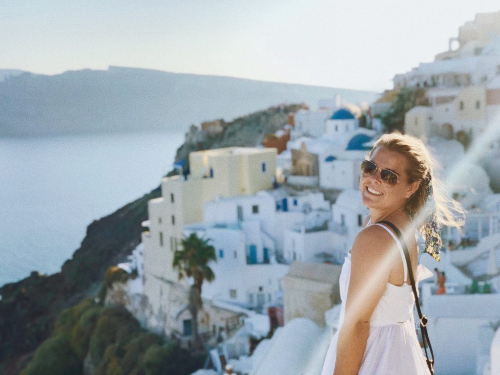 Molly on Santorinini. She is standing in front of the town looking back over her left shoulder toward the camera. She's wearing a white dress with spaghetti straps and sunglasses with her purse over her left shoulder and across her body. The town is in front of her to the left in the photo and the ocean and distant island are visible in the top left of the photo.