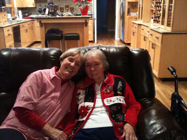 Diane sitting on a black leather couch with her mother. Her mother's kitchen with light wood cabinets can be seen in the background. Diane is on the left wearing a light red collared blouse over a long sleeve red t-shirt. Dian's mom is wearing dark pants, a white shirt and a red and black Christmas sweater.
