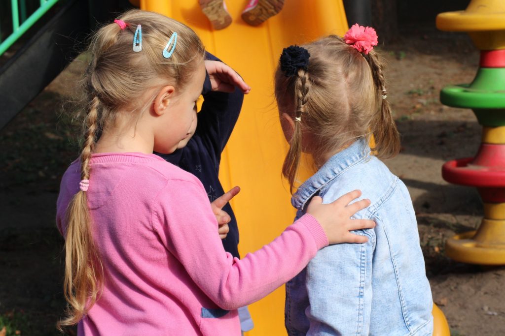 Two little girls in front of a yellow plastic piece of playground equipment. The girl on the left has blond hair with two blue clips in it. She's wearing a long sleeve pink shirt and has one hand on the other little girl's arm, and she's pointing with her left hand. The little girl on the right is wearing a long sleeve blue collared shirt and has her blond hair in two braids