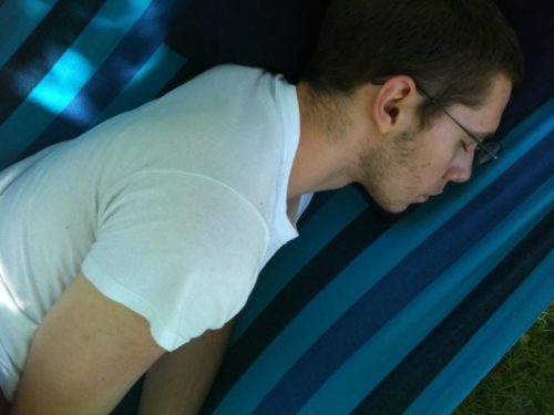 Jordan sleeping on his stomach on a light and dark blue striped hammock. He's wearing wire-rim glasses and a short sleeve white t-shirt. He has dark hair and 3-5 day growth on his cheeks
