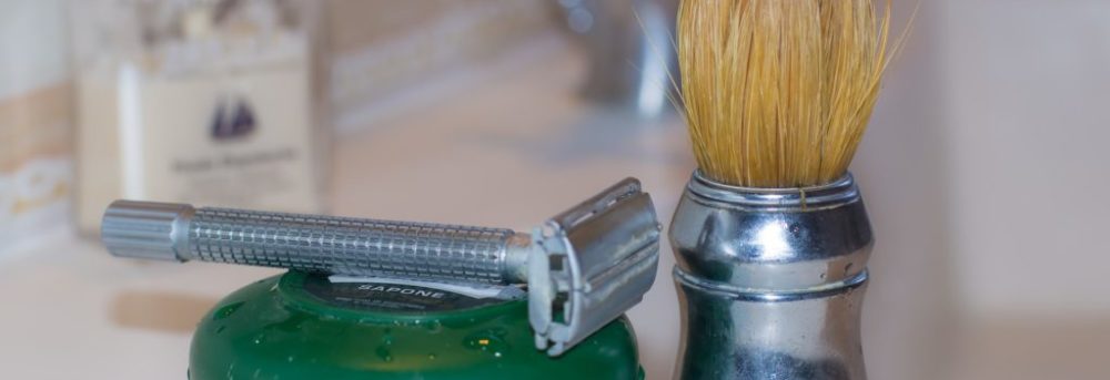 Man's silver razor lying on top of a small green circular container. Sitting next to it is a shaving cream brush with a silver handle