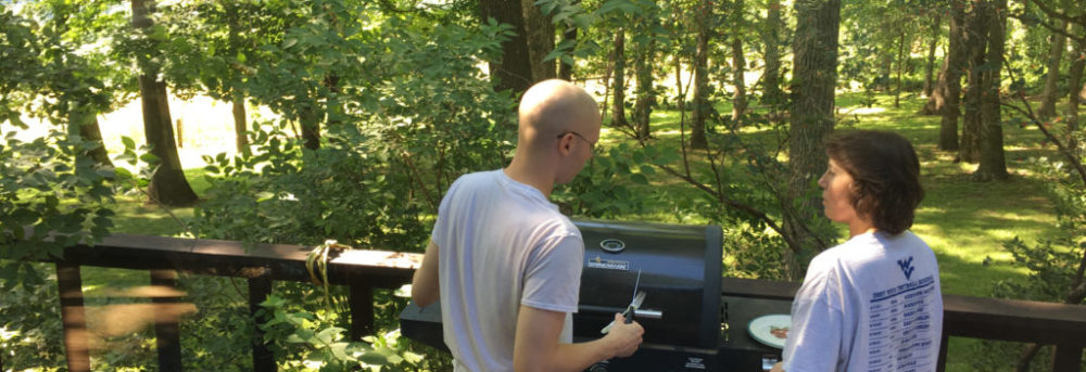 Jordan and Bambi barbecuing on the back deck. Jordan is bald wearing a gray tshirt and black shorts. His back is to us and he's holding a tool. Bambi is on the right wearing a white tshirt with a black shorts. Beyond them is a grove of trees