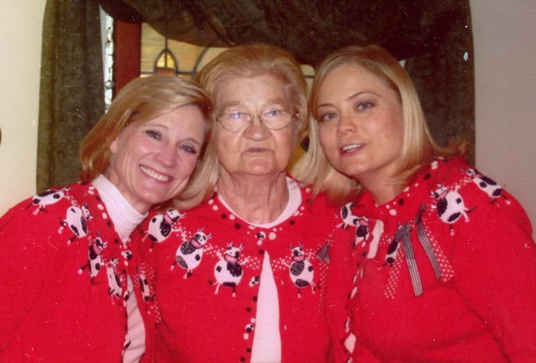 Steph's sister, her mom and Steph. They all have blond hair. Steph's mom, in the middle is wearing glasses. All three are wearing red sweaters with black and white dancing cows just below the collar