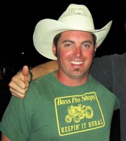 Kyle wearing a white cowboy hat and a green t-shirt that says "Bass Pro Shops Keepin' It Rural" in yellow with a yellow tractor