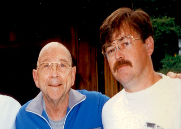 Jerry and his dad. Jerry's dad is bald, wearing glasses, a gray tshirt and a blue jacket with gray color. Jerry is wearing glasses and white t-shirt and has a mustache