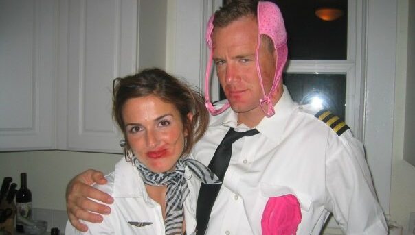 Jen and Matt dressed as a flight attendant and airplane captain. Jen's red lipstick is smeared. She's wearing a white collared blouse and black and white striped scarf tied around her neck. Matt is wearing a white captain's shirt with gold and black epaulets and a black tie. He has a pink bra on his head like ear muffs and pink panties in his left pocket