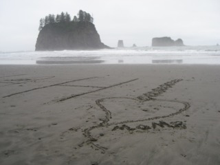 An H and a heart and the word MOM written in the sand on the Oregon coast. A mound with trees on it can be seen rising up in the middle of the water