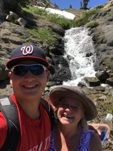 Tommy and Cindy standing in front of a waterfall. Tommy is wearing a navy hat red around the front brim and a script W on it. He's wearing a red t-shirt and a backpack with black straps. Cindy is wearing a tan hat, glasses and a purple shirt