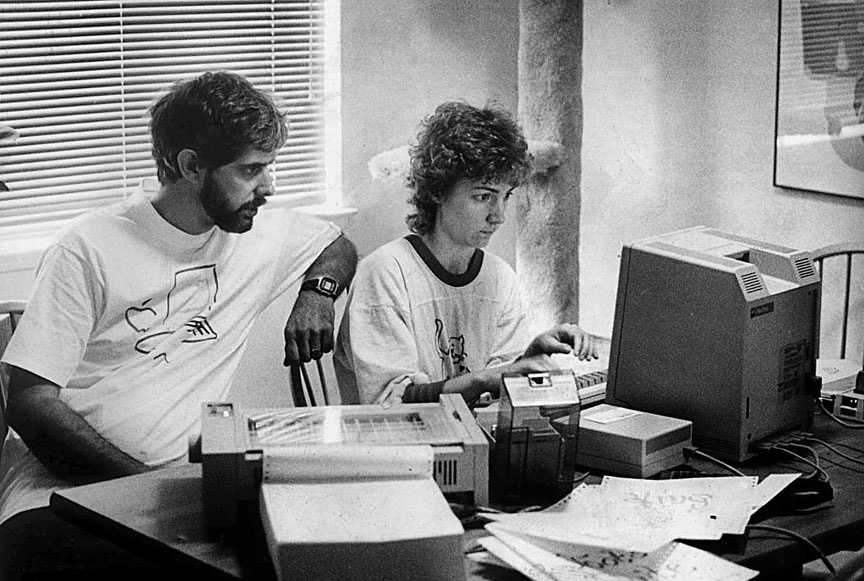 Jan working at her computer with Cliff to her right. The photo is black and white. Cliff has a beard and is wearing a white t-shirt with a logo on it and a watch. He's leaning on Jan's chair. Jan is wearing a white t-shirt with a long sleeve dark t-shirt underneath. They are both seated behind a desk