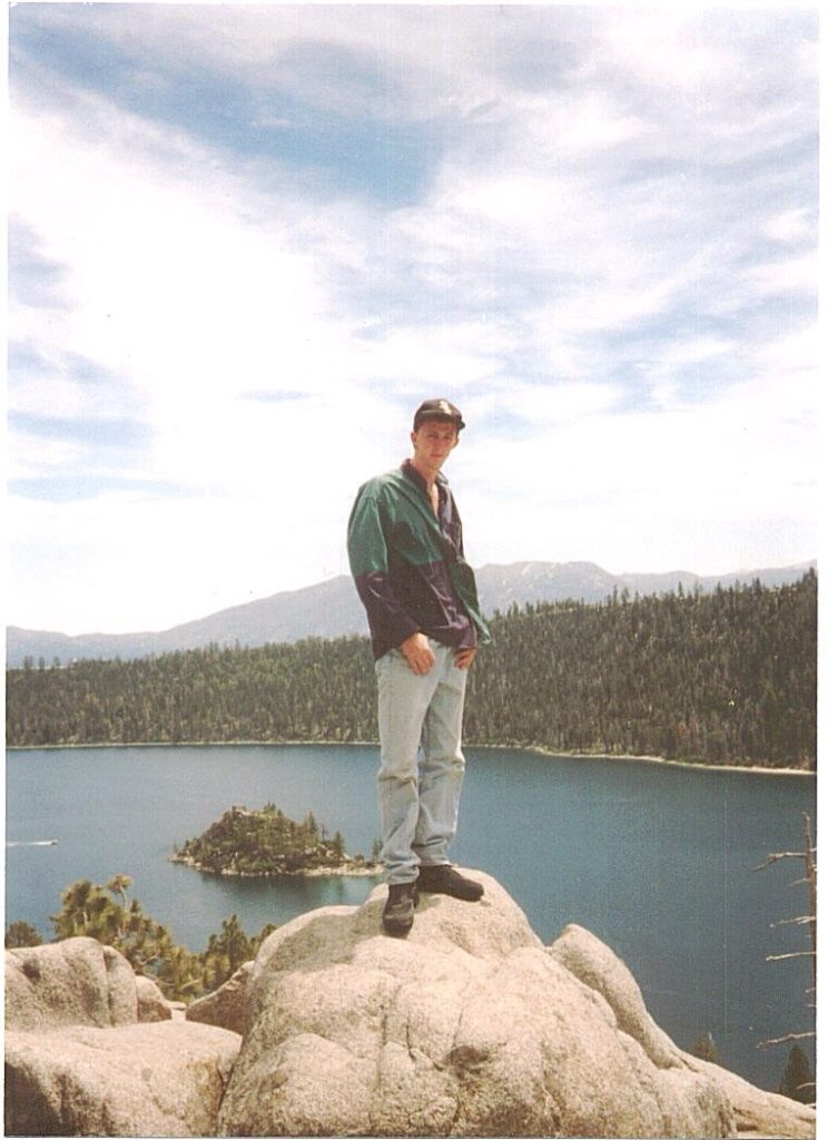 Seth standing on a rock in front of a large lake with pine trees and another mountain in the background. He's wearing light blue jeans, black shoes, a green jacket and a baseball cap