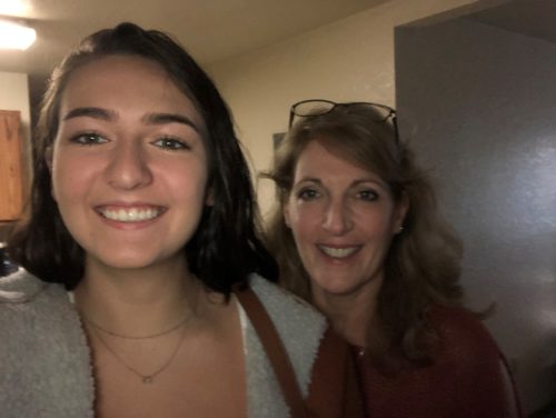 A selfie of Emily and Susan. Emily is on the left wearing a tan coat and has her brown hair parted on the right side. Susan is on the right and has reading glasses on the top of her head. She's wearing a dark red jacket and has shoulder length light brown hair