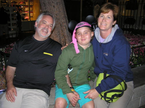 Brian, Jen and MJ at Disneyland. Brian is on the left. He's wearing khaki pants, a long sleeve black LIVESTRONG shirt. He has white gray hair and a beard and mustache. Jenny is wearing a pink hat, olive green jacket and teal shorts. MJ is wearing a blue jacket with a gray color
