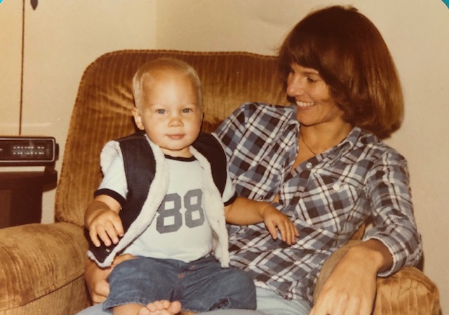 Heidi is sitting on a brown chair. Scott is about two. He's sitting on her lap wearing a dark vest with white trim, a white t-shirt with the number 88 on it and jean pants. His hair is white blond. Heidi's hair is dark brown. She's wearing a black and white checked shirt and light blue jeans. She's looking at him, and he's looking at the camera